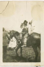 Two young men dressed as Native American caricatures sit on a horse being led by another man.