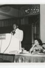 A man stands at a podium at an Alumni Banquet. Written on verso: Clark College Alumni Banquet ca. 1970, Left to right: 1. Scott, 2., 3. James Dean (at podium), 4. Dr. Eula Cohen, 5. Dr. Elias Blake, Jr.