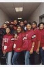 Four young women, wearing matching red sweat shirts, pose with nine men in a hallway.