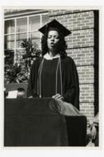 A woman, wearing a graduation cap and gown, stands at the podium on stage at commencement.
