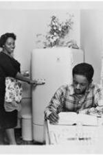 Isaac Mullins studies at a kitchen table as his wife looks on. Written on verso: Gammon Theological Seminary, Student Apartments, Mr. and Mrs. Isaac Mullins.