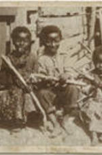 Three children holding sugar cane sit outside of a wooden structure.