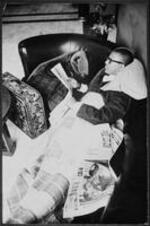 A man relaxes on a couch and reads a newspaper next to his suitcase.