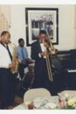 Five men play musical instruments, including drums, saxophone, piano, an upright bass, and a trombone, in a dining room.