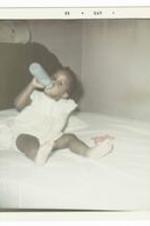 An unidentified baby sits on a bed and drinks from a bottle.