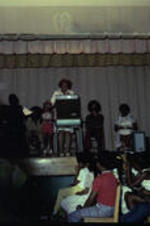 An unidentified woman speaking on stage in front of a crowd of children. Unidentified adults sit in a line in folding chairs.