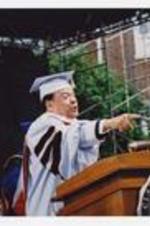 Andrew Young, wearing a graduation cap and gown, stands at the podium on stage at commencement.