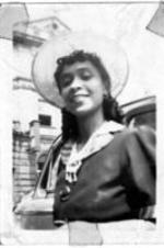 An unidentified woman stands next to a car. She is smiling and wearing a straw hat with a large building behind her.