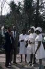 A group of women and men dressed for church stand outside. Unknown location.