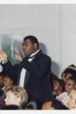 A man, wearing a suit with bowtie, stands in the audience conducting music at convocation.