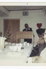 An unidentified man stands behind a podium and talks to an audience seated at a buffet.