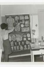 A young woman stands next to a large poster showing parts of human anatomy, in a classroom, a table with science equipment sits off to the side.