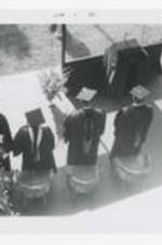 Four graduates wearing caps and gowns, stand on an outdoor stage at commencement.