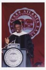A man, wearing graduation cap and gown, stands at the podium at commencement in front of a banner "Clark Atlanta University."