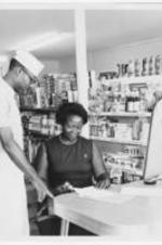 A woman sits and looks over paperwork in a grocery story with a male grocery employee standing beside her.