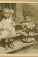 A toddler stands on a house step and waves a flower.
