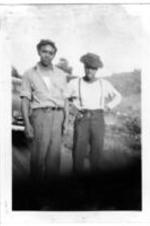 Two unidentified men stand by a car on the side of a rural road.