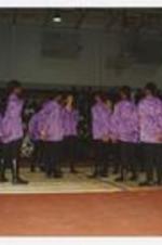 A group of men and women, wearing matching purple jackets, stand on the basketball court at a homecoming step show.