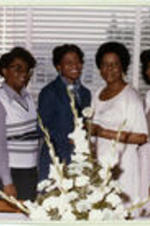 A group of women stand in front of a bouquet of flowers on a table. Written on recto: Hostesses (L-R): Valerie Crawford, Jean Wil Kerson, Franis Mitchell, Mrs. Minnie Wright, Pamela Wyatt, and Christy Esau.
