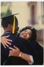 A man and woman wearing graduation caps and gowns embrace at commencement.