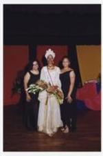 A women, wearing a long-sleeve floor-length white and gold dress with matching head wrap, sash and flower bouquet, poses with two other women on stage.