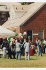 Men and women gather with food on a lawn, next to a brick building, with a pop-up tent and pink and green balloons.