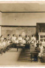 A first grade class sits at desks with their teachers who are normal students.