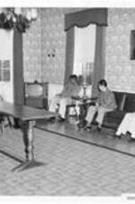 Interior of the dormitory lounge at Ware Hall with students sitting on chairs and couches. Written on recto: Dormitory Lounge-Ware Hall.