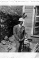 A well-dressed unidentified man stands in front of a house. Next to him is a  stone wall. In the house, another unidentified man is entering the house through a door.