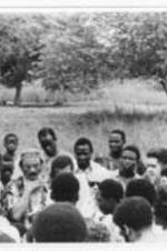 A crowd of people gather in a clearing to listen to a man talk.