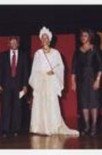 A women, wearing a long-sleeve floor-length white and gold dress with matching head wrap, stands on stage with a man and another woman.