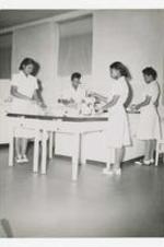 Four women, wearing matching shirt dresses and saddle shoes, stand at work stations with kitchen stand mixers and other kitchen utensils.