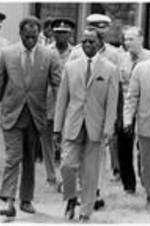 A group of men walk with police in the background. Written on verso: Left to right: Professor J. H. Nketia, Director, Institute of African Studies, Professor Alex Kwapong, Vice Chancellor, University of Ghana, Prime Minister Busia. Second row: Man behind Nketia with dark glasses, Mr. M. Dowvona, Chairman of council for higher education. Two military men. White man, Richard Greenfield, Administrative Secretary, Institute of African Studies.