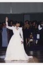 A young woman, wearing a long-sleeve, floor-length dress, stands with her right hand raised in the center of the dance floor, men and women are seated at tables in the background.