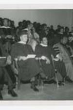 James P. Brawley, C. Eric Lincoln and  other men and women, wearing graduation robes, sit in chairs on stage at commencement.