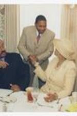 Wynton Marsalis shakes hands with a woman seated between two men at a dining table in a room with floral wallpaper.