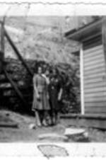 An unidentified group standing outside of a house. Three women stand close together at the bottom of a wood staircase. An unidentified man stands on a porch leaning on a wooden railing.