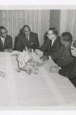 Nine men, wearing suits with neckties, sit around a table with floral arrangement in the center.