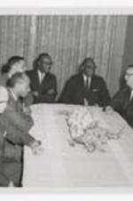 Nine men, wearing suits with neckties, sit around a table with floral arrangement in the center.