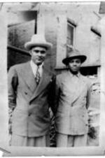 Two unidentified men wearing suits stand in front of a brick building.