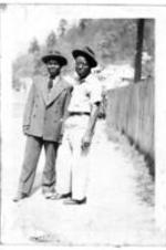 Two unidentified men wearing hats stand next to a fence on a sunny day.