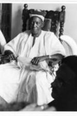An elderly man holding a horse hair fly swatter sits in a wooden chair decorated with elephant tusks. He is wearing white robes that cover him almost completely.