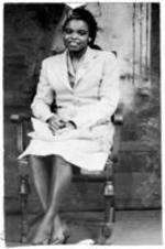 Protrait of an unidentified woman sitting in a chair with her legs crossed in front of a backdrop.
