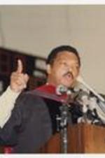 Jesse Jackson, wearing a graduation robe, stands at the podium with multiple microphones at convocation.
