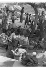 Women and girls working in the shade. Large baskets are stacked along  the ground. Canned and jar goods are sitting on small wooden tables.