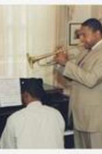 Wynton Marsalis plays his trumpet with a man playing the piano, in a dining room.