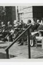 Men and women, wearing graduation caps and gowns, sit on an outdoor stage at commencement.