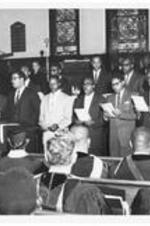 A group of men singing in the campus chapel. Written on verso: (L to R) Front row: Maurice King, Jesse Douglas, Mace Pleasure, (three unidentified), Joseph Epps. Second row: J, Joshua Hutchens, (unidentified), Mr. Stafford Smuting, Mr. Grady Butler.