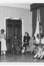 Seven women sit and stand in the Bumstead Hall lounge, while on puts a record on. Written on verso: Dormitory life- resident students relax in the lounge of Bumstead Hall. Fall, 1952