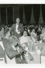 View of man holding paper, standing among men and women seated at dining tables.